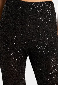 Pantalones de lentejuelas negras con una superficie texturizada, adornada con lentejuelas brillantes y densamente agrupadas, y una cinturilla ajustada. Acabado suave con detalles reflectantes.