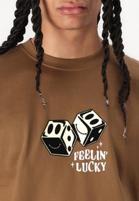 Brun bomulds T-shirt med et grafik af to sorte og hvide terninger med smilende ansigt og teksten "FEELIN' LUCKY" nedenunder.