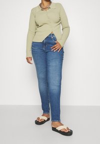Cárdigan de manga larga de color verde claro con canalé y cierre de botón, combinado con jeans ajustados azules y sandalias de plataforma negras con correas crema.