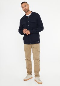 Námořnický pletený kardigan se dvěma kapsami na prsou, kulatým límcem a zapínáním na knoflíky, v kombinaci s béžovými cargo kalhotami a bílými teniskami.