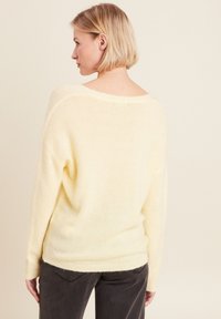 Gelber, übergroßer Pullover aus weichem, flauschigem Material mit weitem Ausschnitt und geripptem Saum, kombiniert mit dunkelgrauen Hosen.