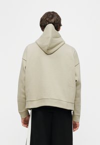 Άνδρας που στέκεται με την πλάτη και φοράει ανοιχτό μπεζ φούτερ με κουκούλα και μαύρο παντελόνι με φαρδιές λευκές κάθετες ρίγες.