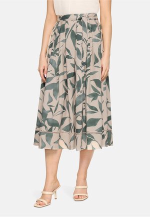 Femme portant une jupe midi beige avec imprimé de feuilles vertes et ceinture nouée à la taille, associée à des sandales à talons hauts beiges.