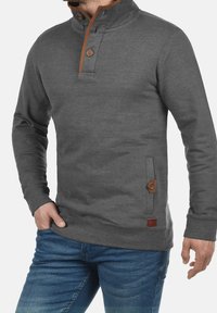 Pullover grigio realizzato in tessuto morbido con colletto alto, dettagli in bottone marrone e tasca laterale con una patch del logo marrone.