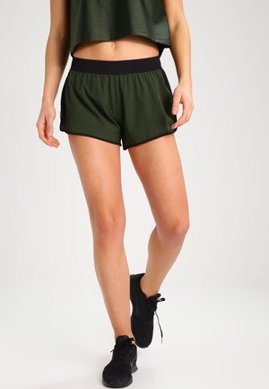 Kvinde iført mørkegrønne træningsshorts med sort linning og sorte sneakers, stående med det ene ben let fremad på hvid baggrund.