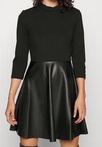 Femme portant un haut noir ajusté à manches trois-quarts et une jupe évasée en cuir noir.