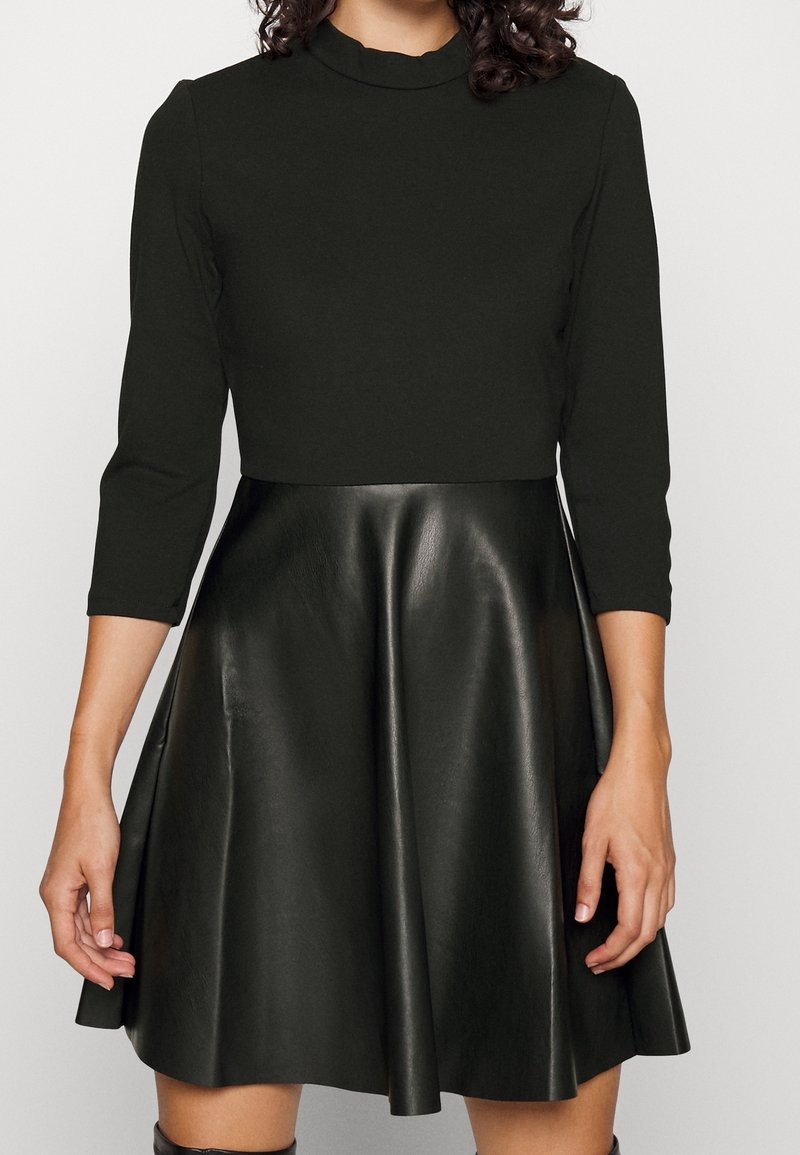 Femme portant un haut noir ajusté à manches trois-quarts et une jupe évasée en cuir noir.