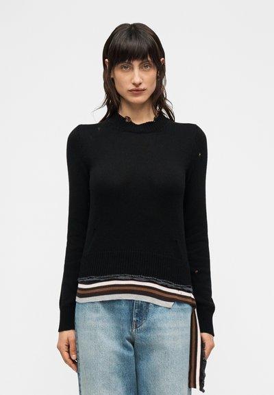 Femme portant un pull noir ajusté avec des détails usés et un ourlet rayé, associé à un jean bleu clair délavé, debout devant un fond uni.
