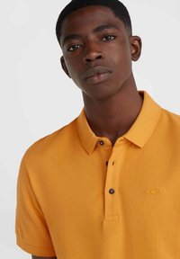 Polo de color mostaza con un acabado texturizado, que presenta una tapeta de tres botones y botones en azul que contrastan. Bordado sutil del logo.