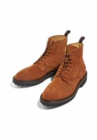 Stivali alla caviglia in suede marrone con un design brogue, caratterizzati da intricate perforazioni, lacci marroni e suola in gomma per una buona aderenza.