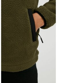 Pull-over en polaire verte avec une surface texturée, des accents noirs et une poche sur le côté avec un détail de cordon de serrage, adapté pour un look décontracté.