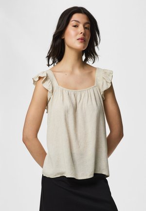 Femme aux cheveux foncés portant un top beige sans manches à volants et une jupe noire, posant les mains derrière le dos devant un fond uni.