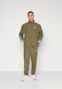 Φόρμα ελιάς πράσινου χρώματος με λευκές ρίγες στο πλάι και ψηλό γιακά. Διαθέτει κλείσιμο με φερμουάρ, ελαστικές μανσέτες και λογότυπα στο στήθος και το παντελόνι.