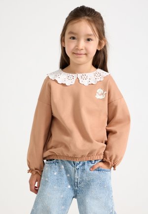 Ung pige med langt hår bærer en tan sweatshirt med hvid blondekrave og katte-patch, kombineret med lyseblå denimjeans med stjernemønster.