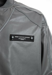 Chaqueta de cuero gris con material texturizado, costuras curvadas y un parche de etiqueta con acentos negros y dos botones a presión.