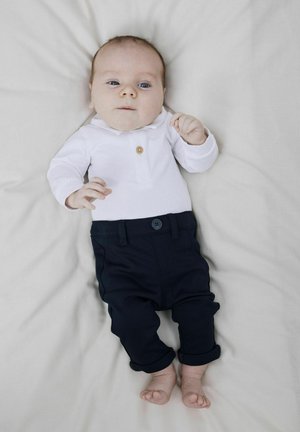 Polo bianco a maniche lunghe con un bottone, abbinato a pantaloni blu navy con risvolto. L'infante giace su uno sfondo chiaro.