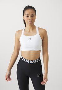 Hvit athletic crop-top laget av elastisk materiale, med en svart Under Armour-logo. Sammen med svarte leggings og en grå linning.