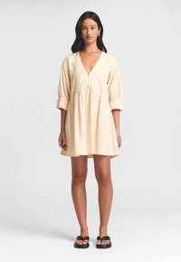 Helles beige kurzes Kleid mit V-Ausschnitt, Ballonärmeln und gerafftem Bund. Der Stoff ist glatt, mit Knöpfen am Ausschnitt, kombiniert mit schwarzen Sandalen.