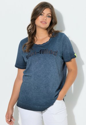 Mujer con cabello largo y castaño que lleva una camiseta azul «llena de sol» y pantalones blancos, de pie con una mano en el bolsillo frente a un fondo claro.