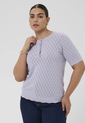 Frau trägt ein lavendelfarbenes kurzärmeliges Strickoberteil mit Diamantmuster und Knöpfen, kombiniert mit dunkelblauen Jeans und goldigem Schmuck.