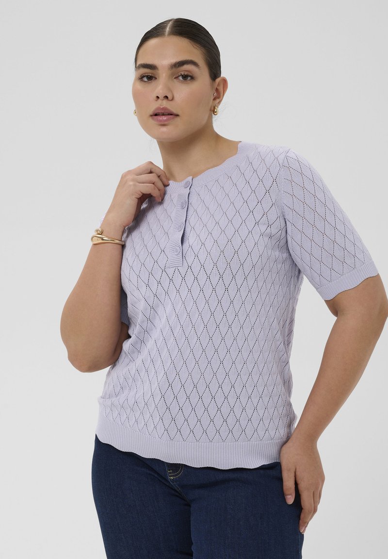 Femme portant un haut en tricot lavande à manches courtes avec un motif en losanges et des boutons, associé à un jean bleu foncé et des bijoux en or.