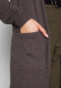 Pletený hnědý cardigan s přední kapsou, žebrovanými manžetami a měkkou texturou. Oblečení má volný střih a otevřený přední design.