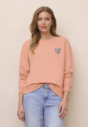 Femme portant un sweat-shirt pêche avec un cœur imprimé léopard sur la poitrine et un jean taille haute bleu clair, debout devant un fond beige.