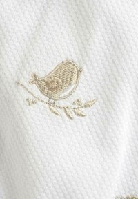 Un tessuto bianco presenta un uccello ricamato in beige chiaro, posato su un ramo con foglie, che enfatizza la texture e i dettagli del ricamo.