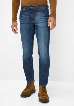 Denim jeans in medium blue with a straight leg, five-pocket design, tan stitching, and a button closure. Worn with brown suede boots.