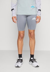 Shorts de sport gris au design ajusté, fabriqués en tissu extensible. Logo blanc sur le côté gauche, portés avec un haut à manches longues et des baskets.