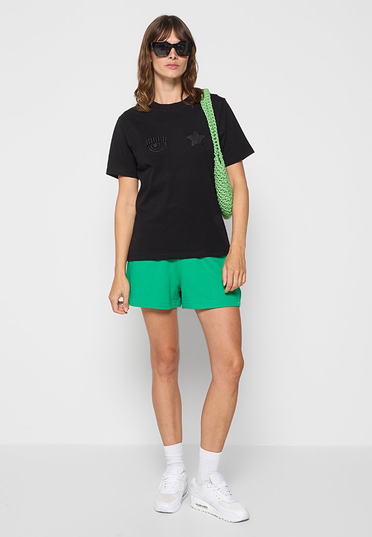 Czarny T-shirt z wzorami oka i gwiazdy, zestawiony z zielonymi szortami. Ubranie jest uzupełnione zieloną plecioną torbą i białymi sneakersami.