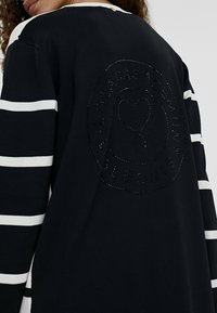 Felpa navy con righe orizzontali bianche. Presenta un'emblema rotondo e testurizzato con un design a forma di cuore e dettagli scintillanti sul retro.