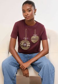 Burgundia T-särk, millel on kuldse tikandiga ornamentaalsed disainid, sisaldades paelu ja keerulisi mustreid. Kombineeritud helesiniste laiade pükstega.