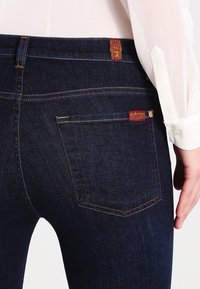 Detailní záběr osoby, která nosí tmavě modré džíny s viditelnou zadní kapsou a červenými látkovými značkovými štítky, doplněné bílou košilí.