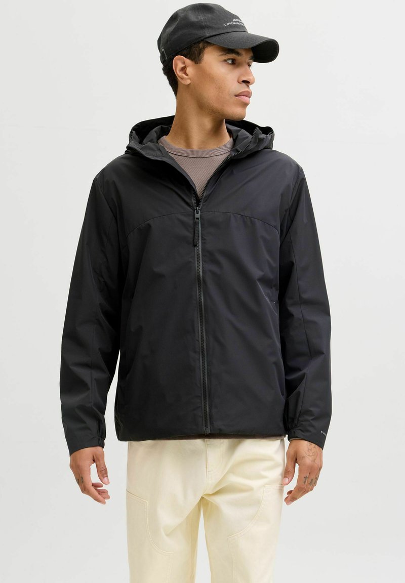 Veste noire légère avec capuche, fermeture éclair et texture lisse. Portée sur un haut marron et associée à un pantalon de couleur claire.