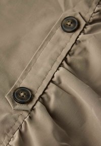 Bežna tkanina s naboranom teksturom, koja sadrži dva tamna dugmeta i šivenu šav uz rub. Prikaz u krupnom planu naglašava detalje.