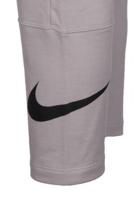 Pantalons de survêtement gris clair avec un logo Swoosh noir de Nike sur la cuisse gauche, fabriqués à partir d'un tissu doux et texturé avec des détails de couture visibles.