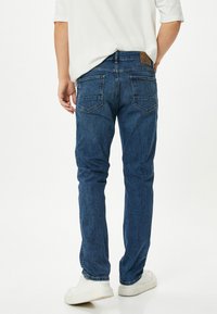 Jeans de mezclilla azul oscuro, corte recto, con dos bolsillos traseros con costura decorativa, parche de cuero en la cinturilla, combinados con zapatillas blancas.
