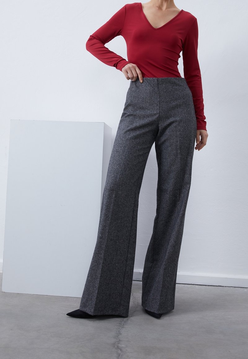 Rotes Langarmoberteil mit V-Ausschnitt, kombiniert mit hoch taillierten, weit geschnittenen grauen Hosen aus einem strukturierten Stoff. Schwarze spitze Schuhe runden den Look ab.