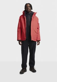 Red waterproof jacket with a high collar, front pockets, and a zipper, worn over a black hoodie and paired with navy pants and boots.