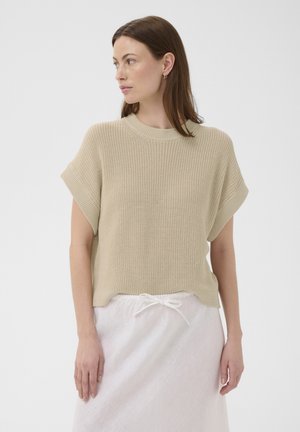 Femme portant un pull en maille beige à manches courtes et une jupe blanche avec cordon, regardant vers sa gauche sur un fond blanc uni.