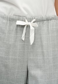 Pantalon à carreaux gris avec une ceinture élastique et un nœud en tissu blanc au centre devant, porté avec un top blanc.