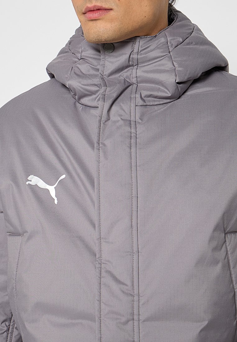 Chaqueta acolchada gris claro con un cuello alto, que cuenta con un cierre de cremallera y un logo blanco en el pecho. Tejido texturizado con un patrón sutil.