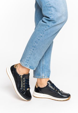 Person trägt glänzende schwarze Sneaker mit goldenen Akzenten und weißen Sohlen, kombiniert mit hellblauen Jeans vor weißem Hintergrund.