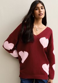 Burgundy knitted sweater featuring pink heart motifs, V-neck, and loose fit with ribbed hem. Worn with denim.