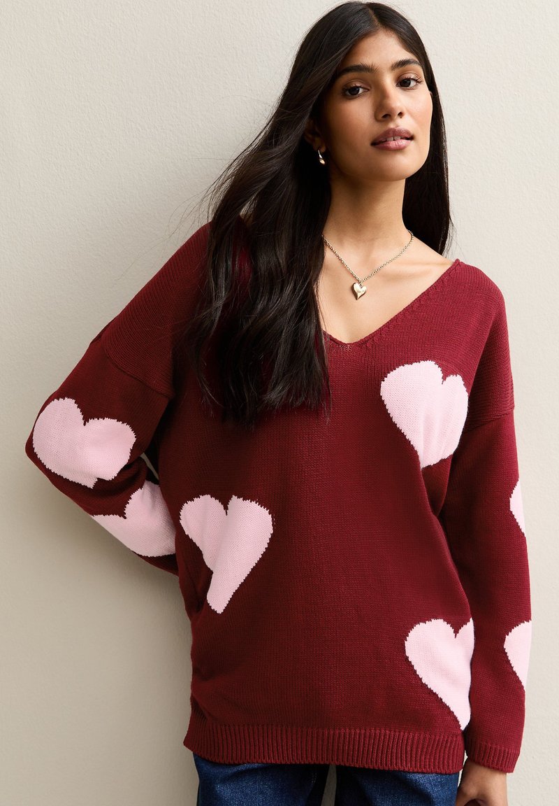 Burgundy knitted sweater featuring pink heart motifs, V-neck, and loose fit with ribbed hem. Worn with denim.