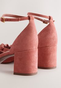 Paire de sandales à talons blocs en daim rose poudré avec brides à la cheville et boucles dorées sur un fond neutre.