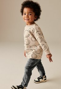 Hellbeige Sweatshirt mit schwarzen Smiley-Gesicht-Mustern, kombiniert mit dunkelgrauen Jogginghosen und schwarzen Sneakers. Das Kind lächelt und läuft.