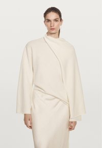 Haut en maille douce couleur crème avec manches longues et un design cache-cœur asymétrique. Présente une texture subtile et un drapé pour une silhouette élégante.