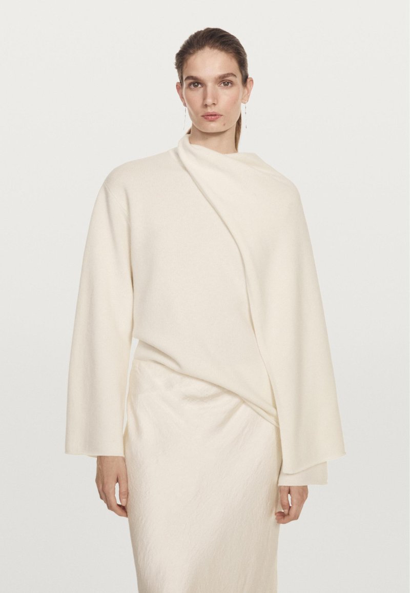 Haut en maille douce couleur crème avec manches longues et un design cache-cœur asymétrique. Présente une texture subtile et un drapé pour une silhouette élégante.